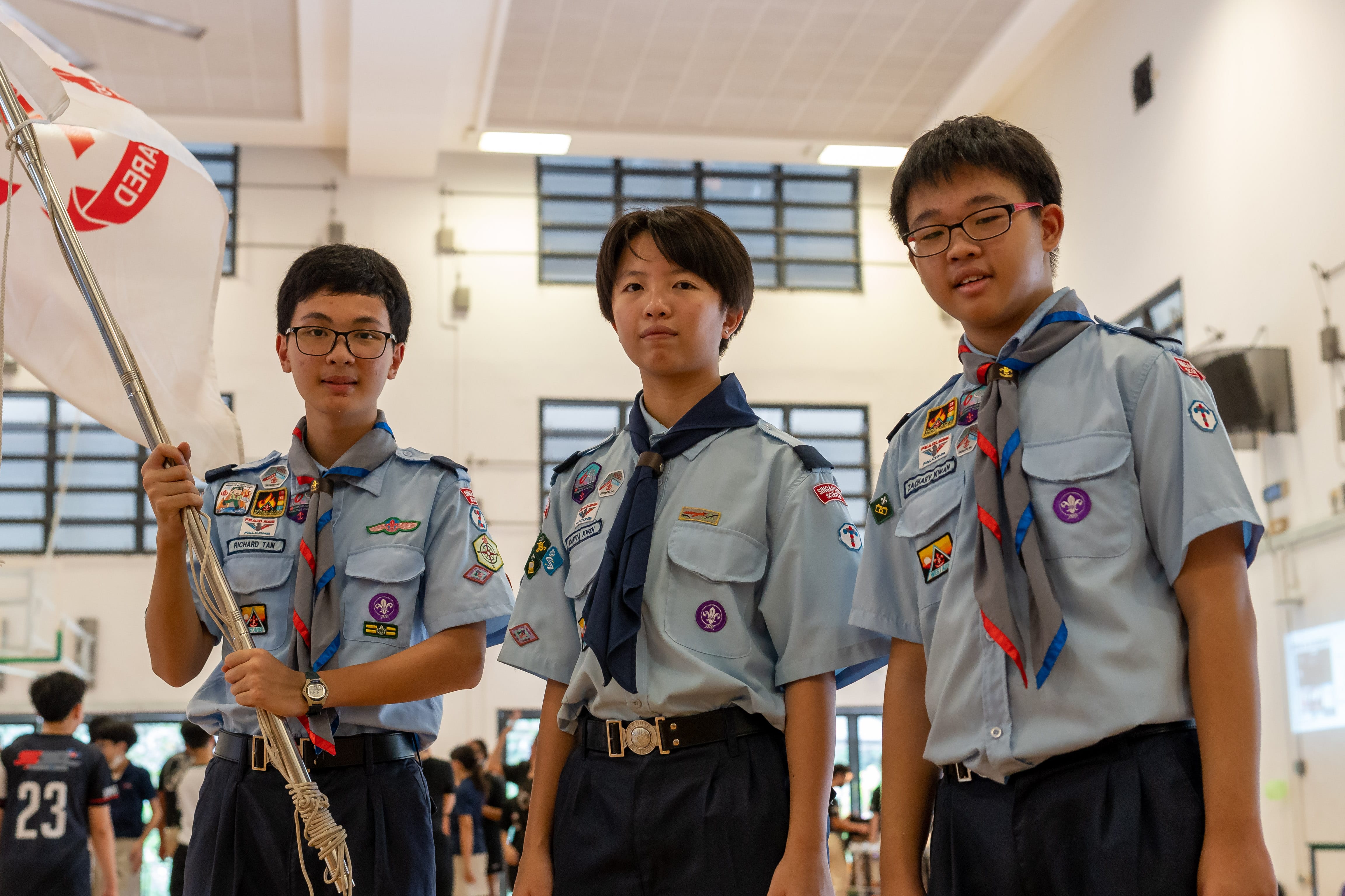 Three young scouts in gray uniforms with badges stand indoors, one holding a white flag with a red emblem.